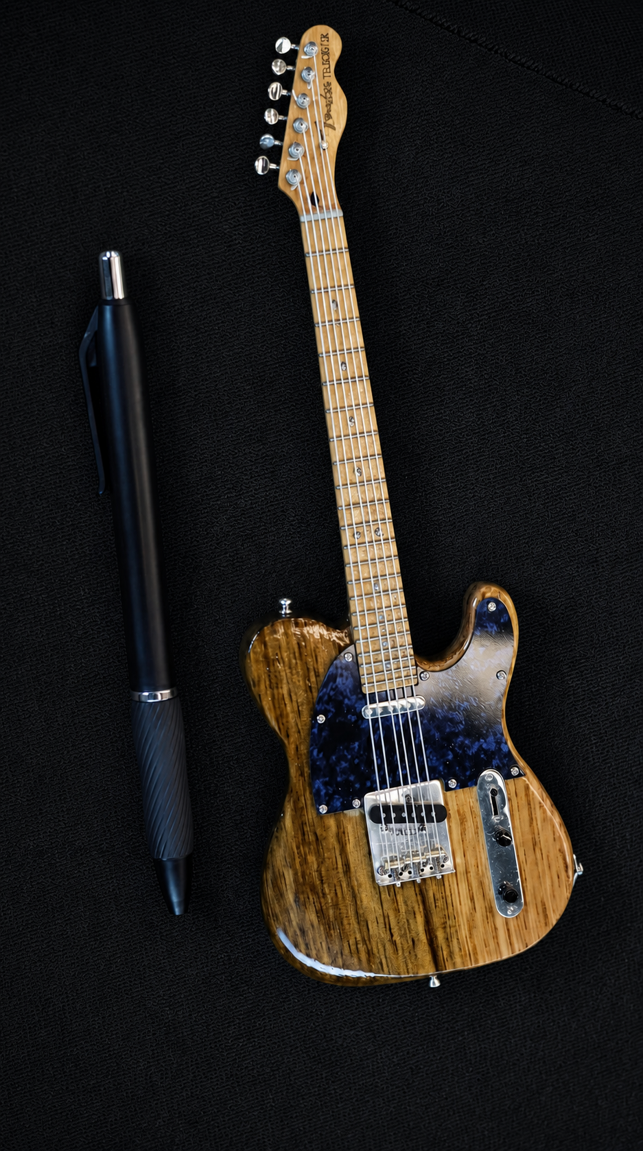 Handcrafted miniature Fender Telecaster replica in California-sourced hardwood with a blue imitation marble plastic pickguard, next to a pen for scale, by Rob Gwynne