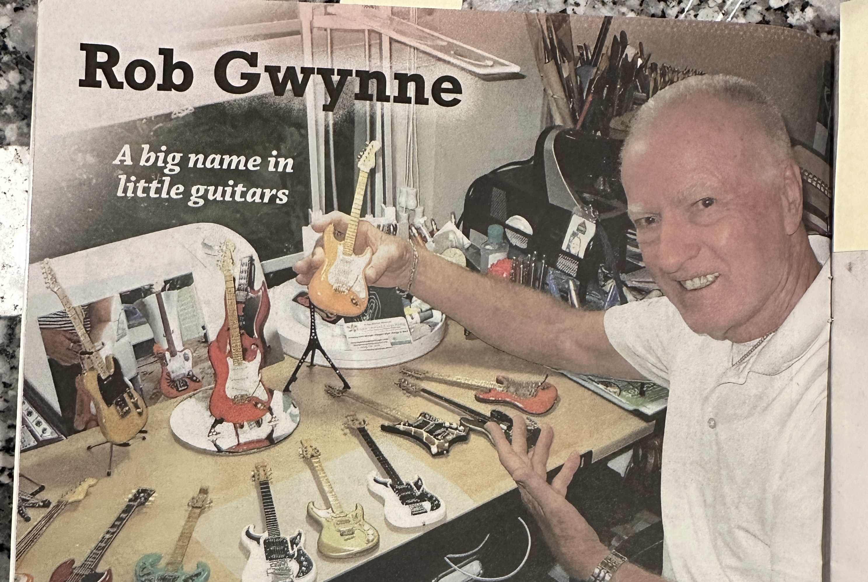 Rob Gwynn smiling at his workbench while holding a finished miniature guitar, with other miniature guitars on the table